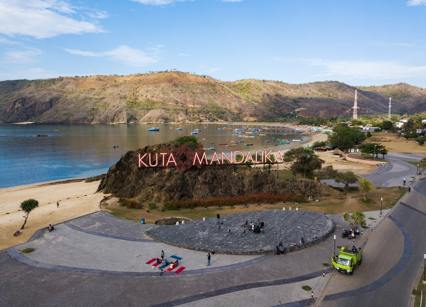 Aerial view of dry coastal hills beside bright turquoise water and a sign promoting Kuta Mandalika.