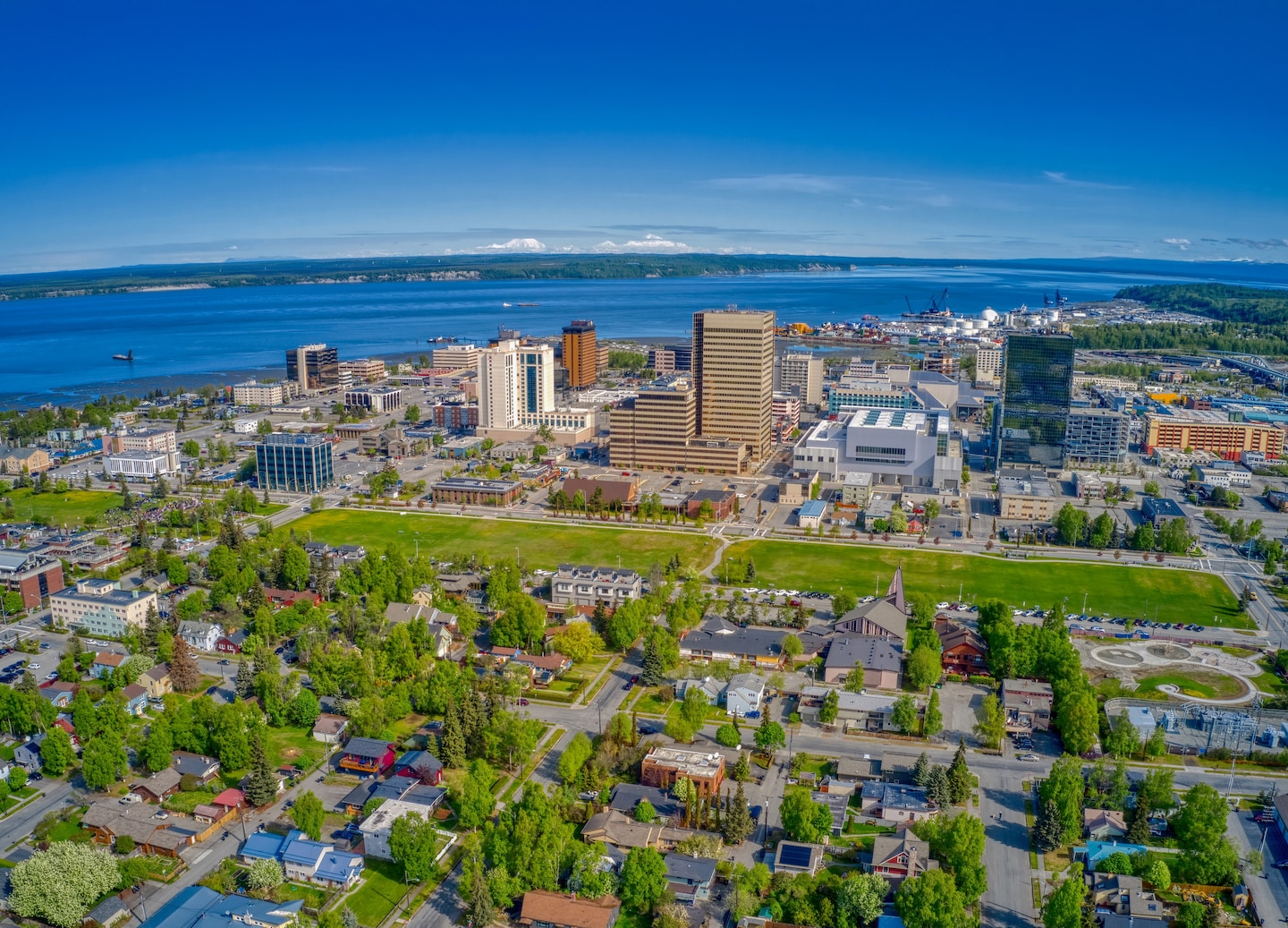 Anchorage’s downtown sits below snow‑covered mountains, with roads and buildings visible from an aerial view. - Anchorage, Alaska