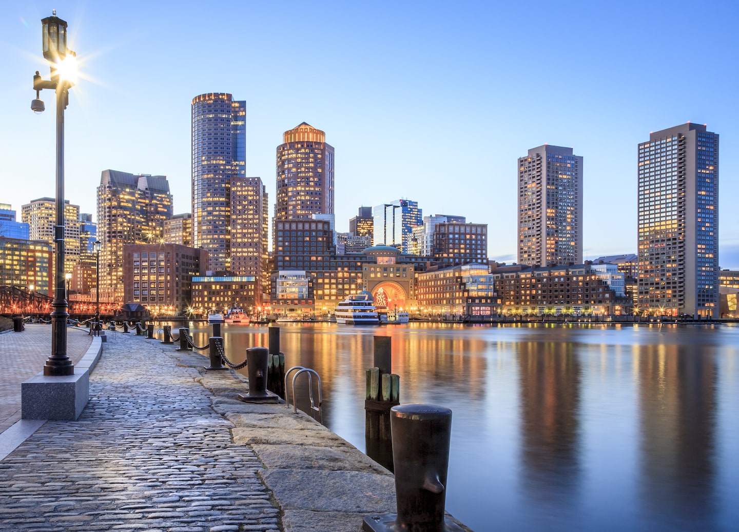Boston Harbor glows at night, with city lights reflecting across calm water beneath the skyline. - Boston, Massachusetts