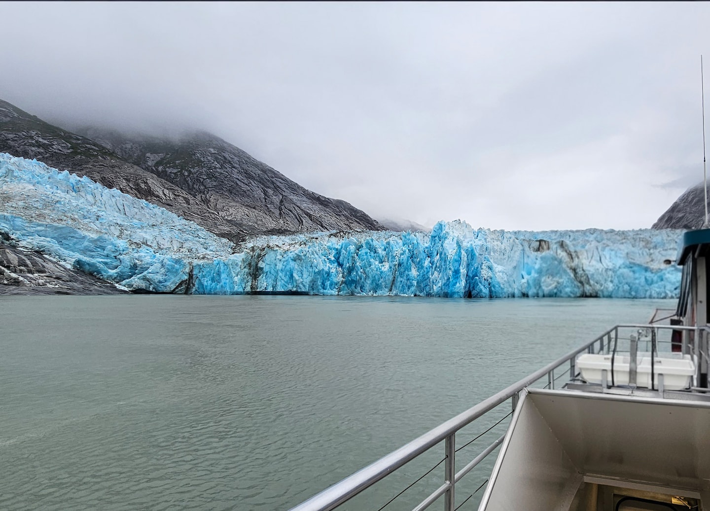 A tour boat approaches Hubbard Glacier’s towering blue ice wall, surrounded by cold, misty Alaskan water. - Hubbard Glacier, Alaska