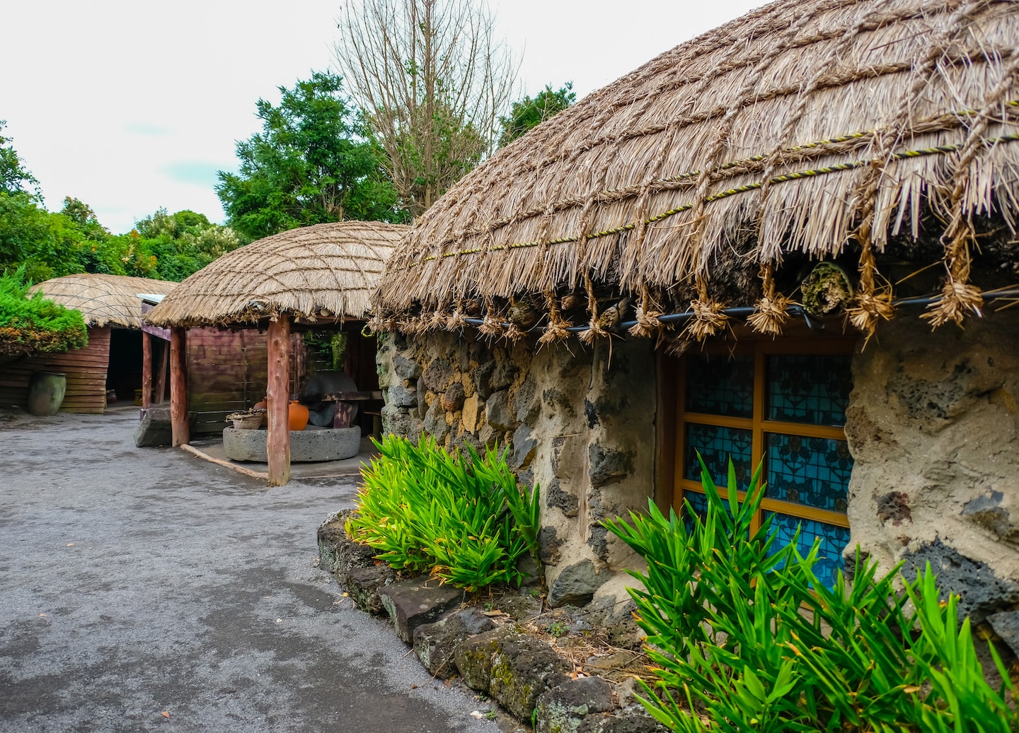 Traditional stone homes line a quiet path in Folk Village, surrounded by greenery and sunlight. - Jeju, South Korea