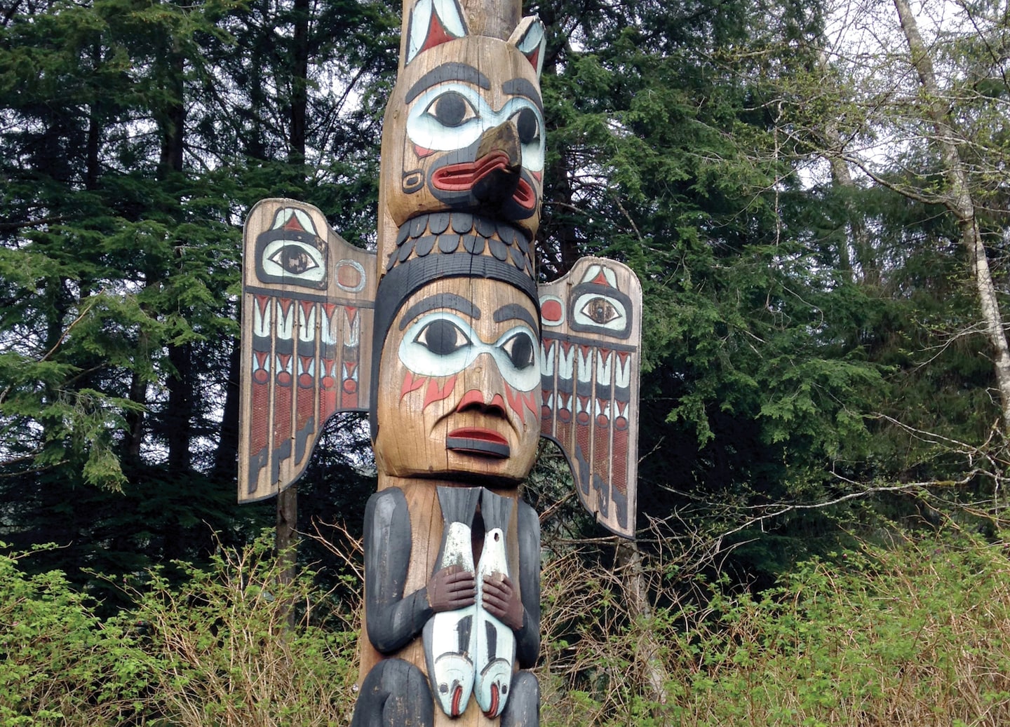 Totem pole rises among forest trees with intricate carved figures. - Ketchikan, Alaska