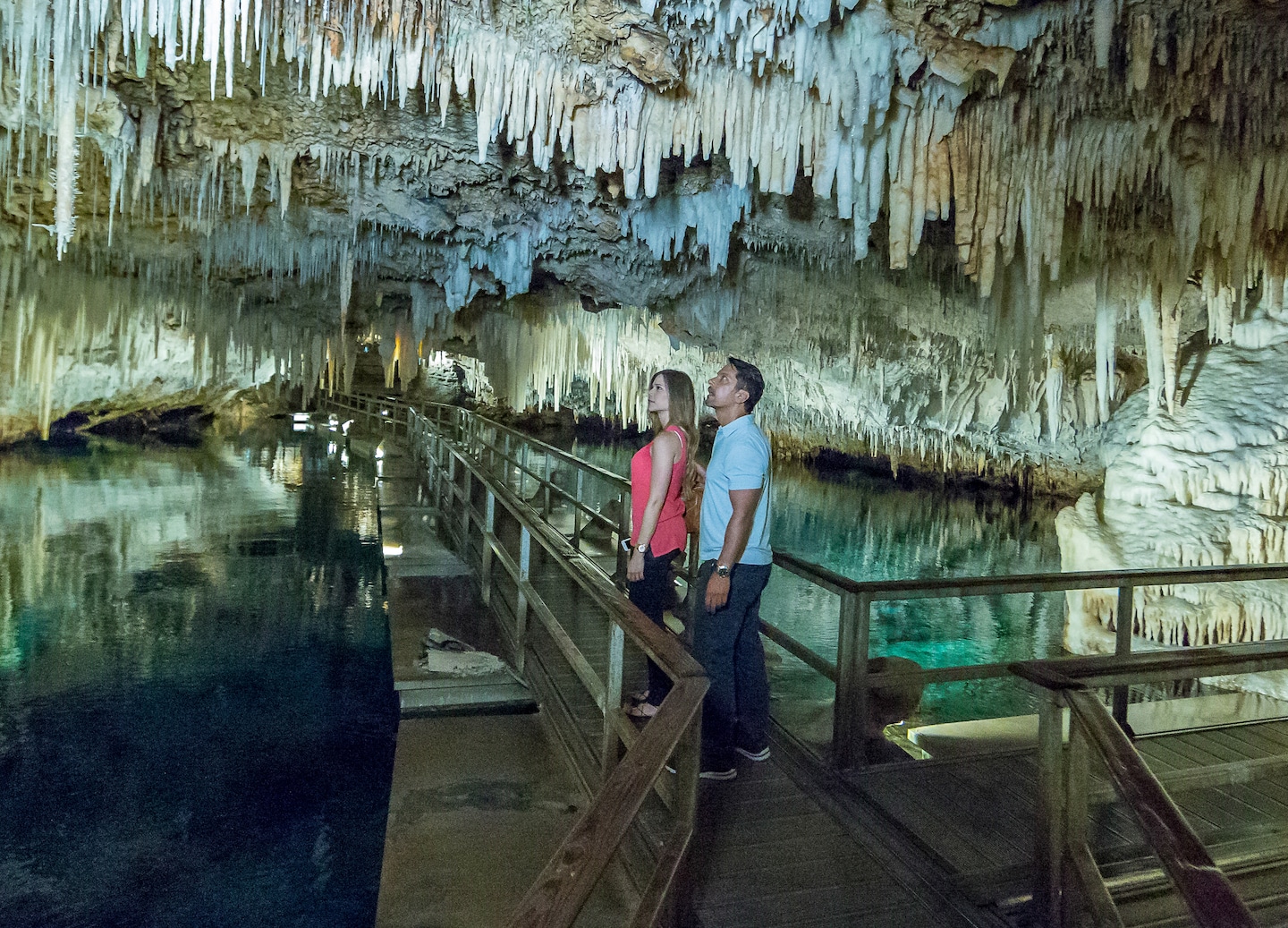 A couple explores a crystal cave walkway surrounded by glowing rock formations and still underground water. - Royal Naval Dockyard, Bermuda