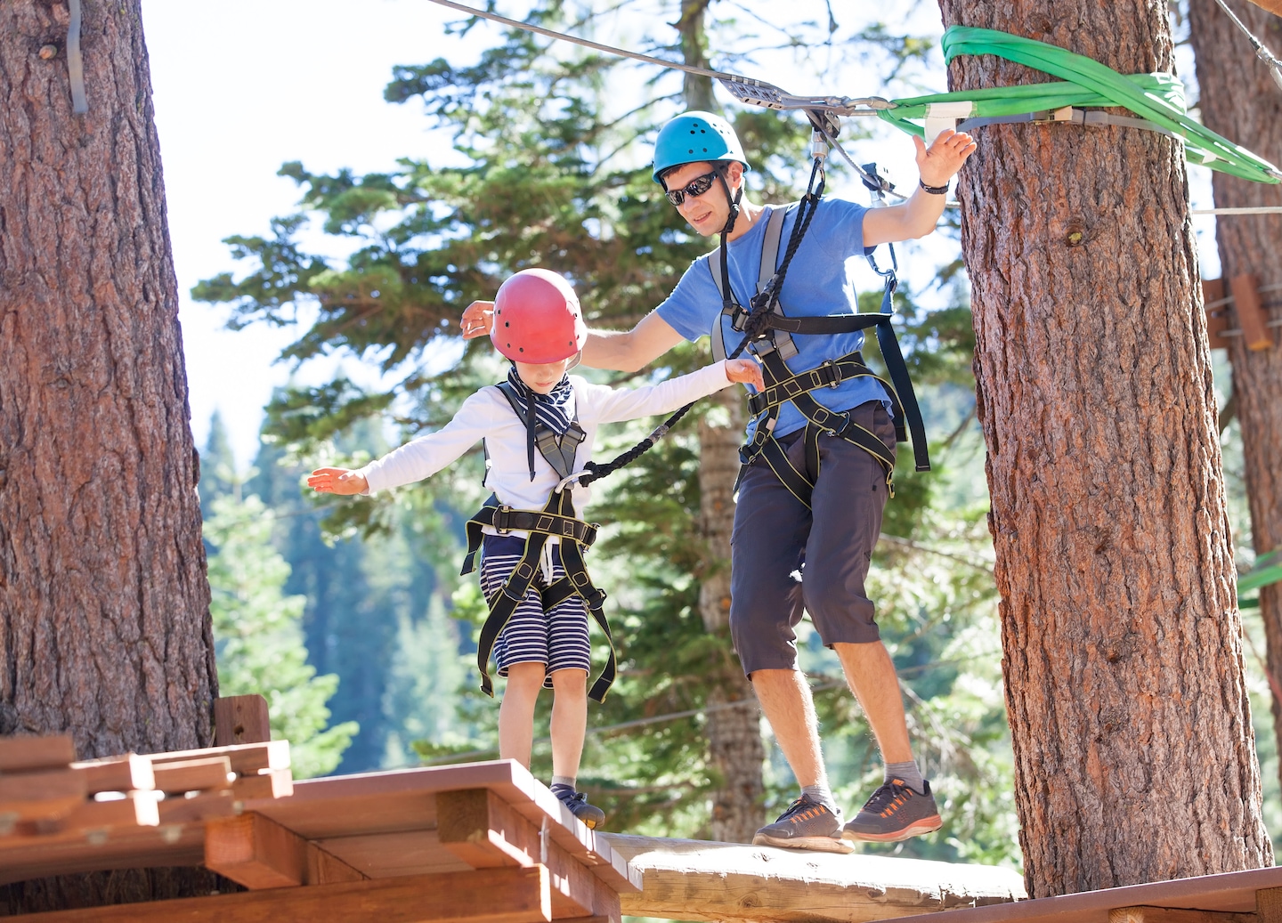 A father and son balance on a ropes‑course beam, holding safety lines above a forested backdrop. - Noumea, New Caledonia