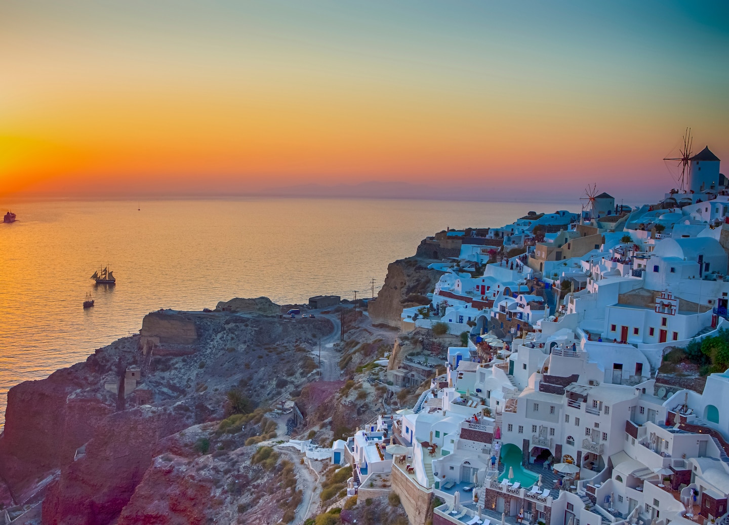 A sailboat glides across Santorini’s caldera as the sun sets behind the cliffs. - Fira, Santorini