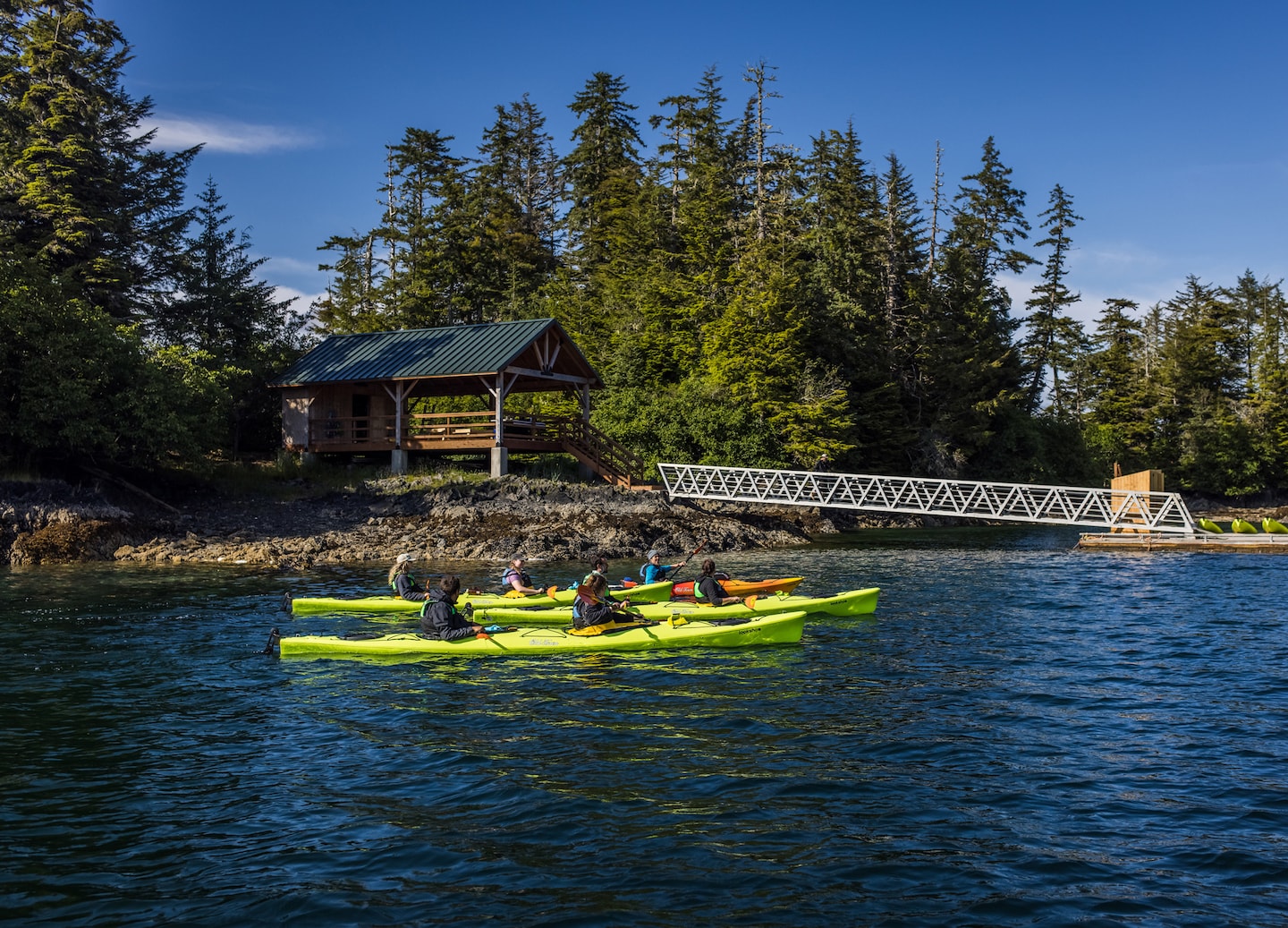 Kayakers approach wooden bridge as calm Siginaka waters reflect surrounding forest. - Sitka, Alaska