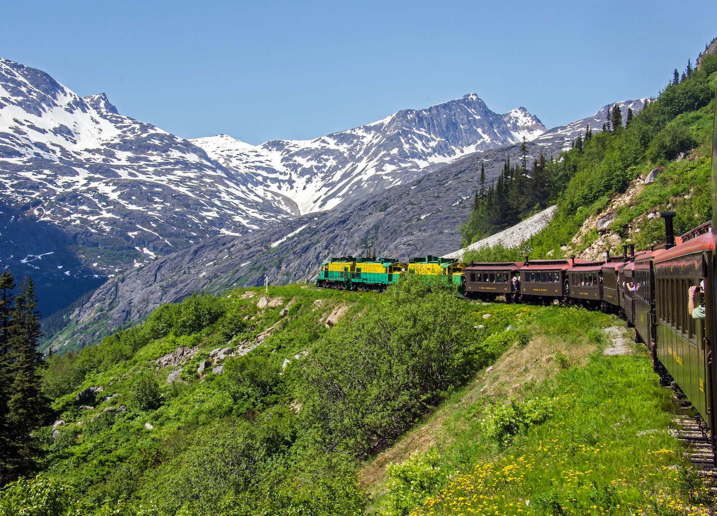 A White Pass train heads along a mountain ridge, surrounded by pine forests and steep slopes. - Skagway, Alaska